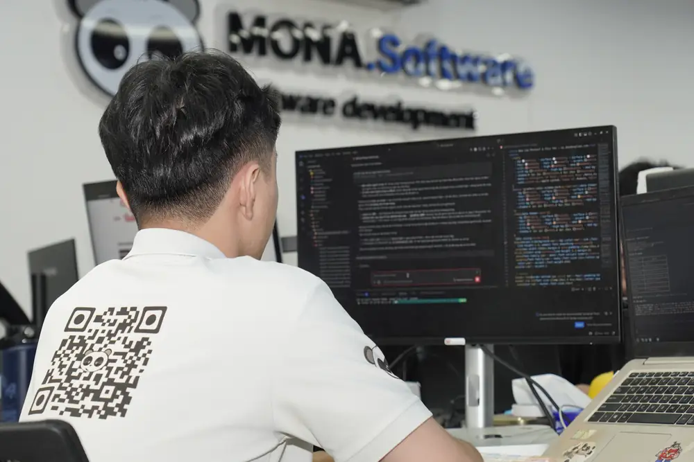 Lập trình viên MONA Software làm việc trước màn hình code, phía sau là bảng logo MONA.Software